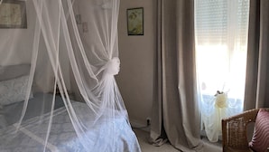 Un dormitorio con cama, ventana con cortinas, mesita de noche y un cuadro en la pared.