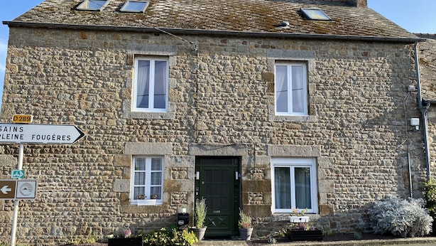 Edificio de piedra con puerta verde, dos ventanas y un cartel que indica direcciones a Sains Pleine y Fougeres.