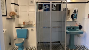 Baño con inodoro azul, cabina de ducha con mampara de vidrio, lavamanos y baldosas de suelo estampadas.