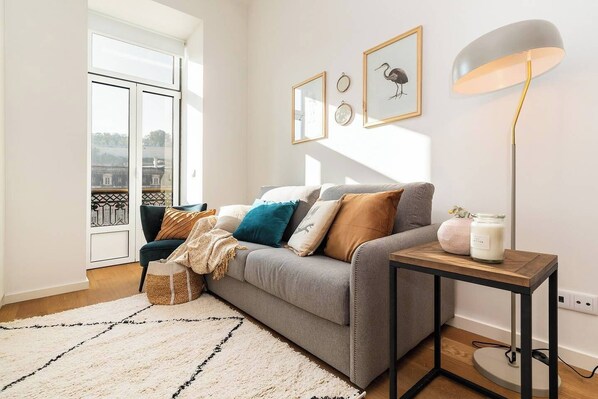 A modern living room with a grey sofa, a wooden side table, and a floor lamp. There are framed pictures on the wall and a view of the city through the window.