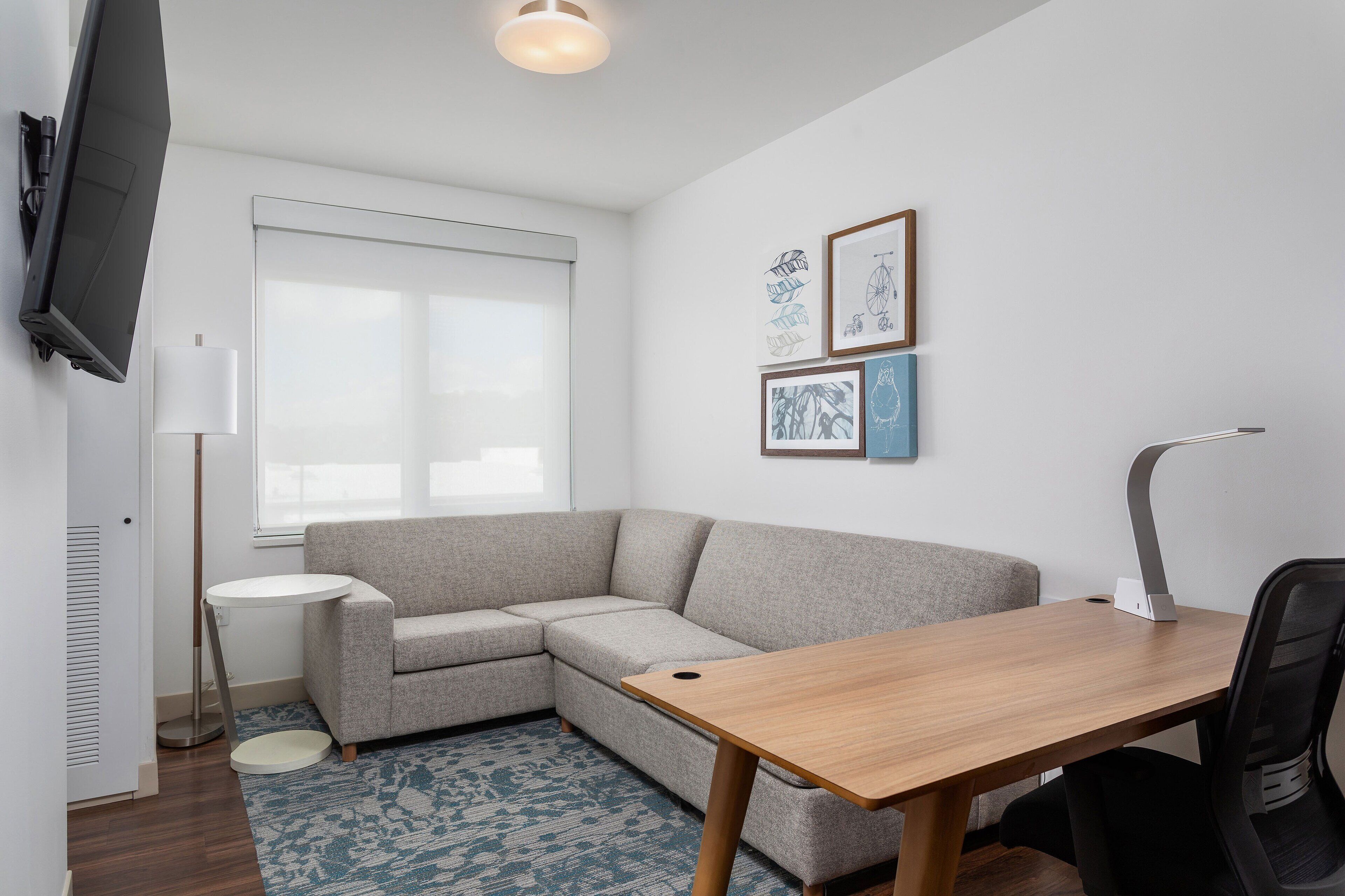 A modern living room with a grey sectional sofa, a wooden dining table, a black office chair, a floor lamp, and a flat-screen TV mounted on the wall.