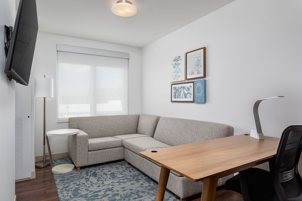 A modern living room with a grey sectional sofa, a wooden dining table, a black office chair, a floor lamp, and a flat-screen TV mounted on the wall.