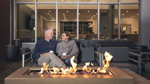 A couple sitting on a couch near a fire feature in a modern indoor setting with large windows and a visible kitchen area in the background.