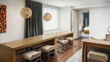 Una habitación de hotel moderna con un escritorio de madera largo, cuatro taburetes altos, un ventanal con cortinas y dos lámparas colgantes esféricas, con vista a la habitación contigua.