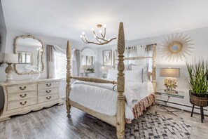 Ein Schlafzimmer mit einem Himmelbett mit Holzrahmen, einer Kandelaberleuchte, einem großen Spiegel und einer Wanddekoration in Form eines Sonnenstrahls.