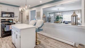 A modern kitchen with white cabinetry, a built-in microwave, and a stove. There is a window with a view of a dining area and a plant. - Charming Ranch on the Cape (Barnstable)