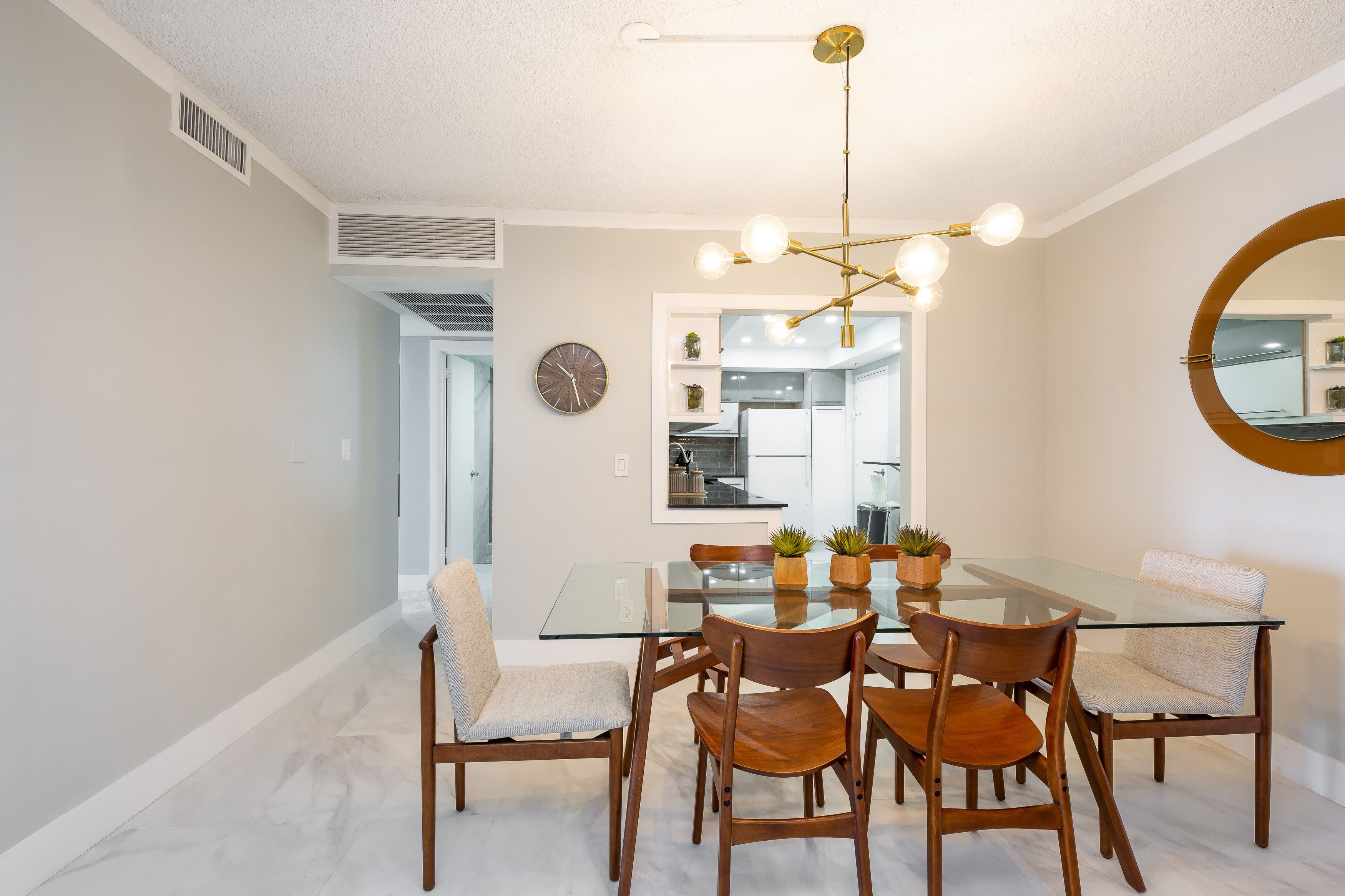 A dining area with a glass table, wooden chairs, a modern chandelier, and a kitchen in the background.
