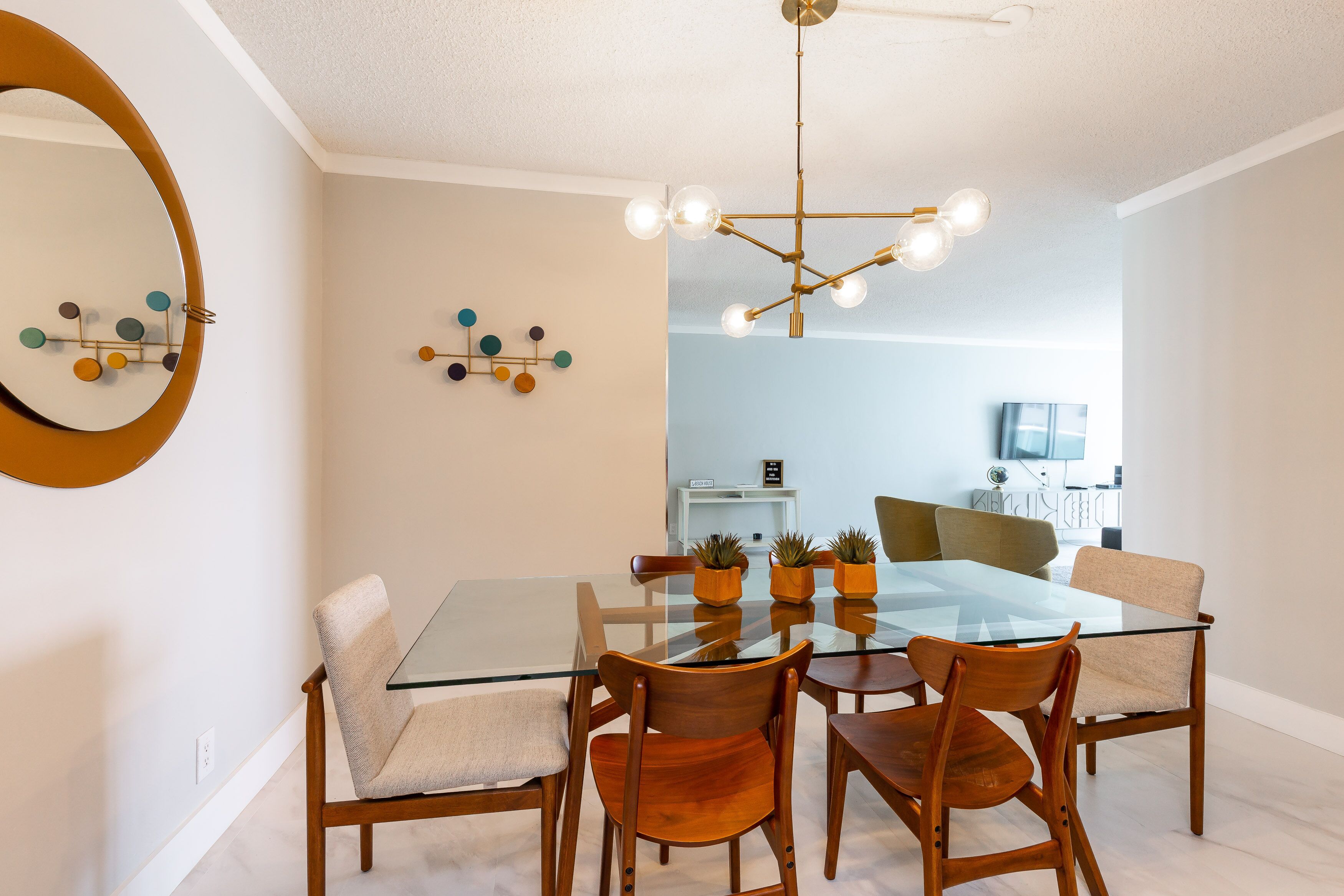 A dining area with a glass table, wooden chairs, a modern chandelier, and a kitchen in the background.