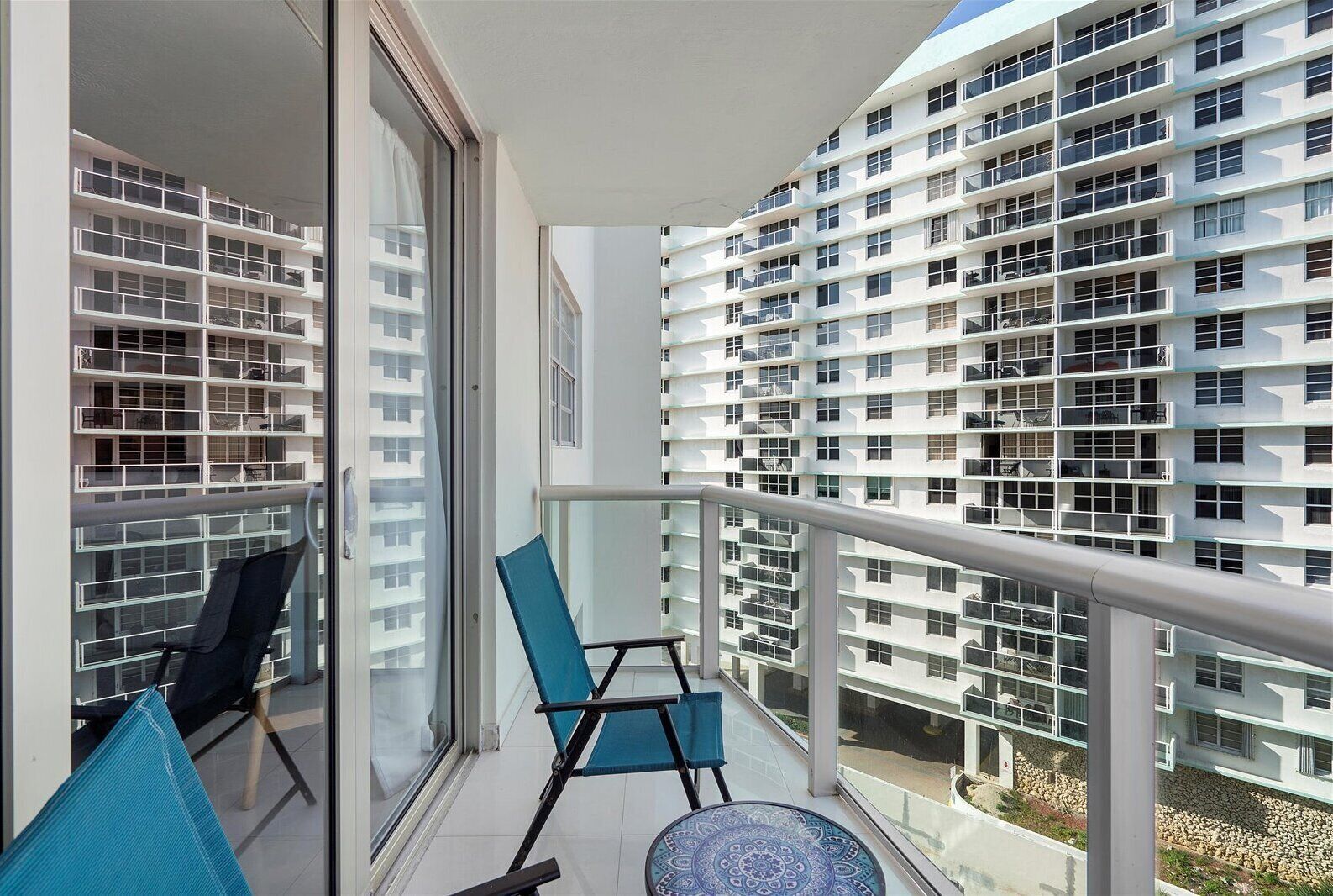 A balcony with a view of a modern building with multiple balconies. There are two chairs and a small table on the balcony.