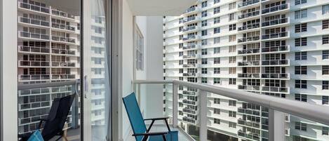 A balcony with a view of a modern building with multiple balconies. There are two chairs and a small table on the balcony.