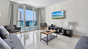 A modern living room with a large TV, a coffee table, and a sofa. There is a balcony with a view of the ocean and buildings. - The Tides Ocean Luxury Suites (Hollywood)