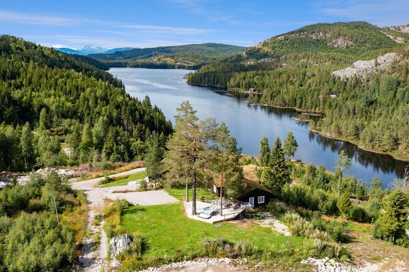 Una cabaña junto al lago con un sendero de tierra que lleva a una terraza, rodeada de un bosque denso y montañas al fondo.