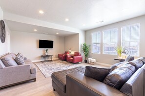 A modern living room with a grey sofa, a brown leather couch, and a red armchair. There is a flat-screen TV mounted on the wall, a small table with a lamp, and a potted plant. - Sun Haven! Pet Friendly! (Washington)