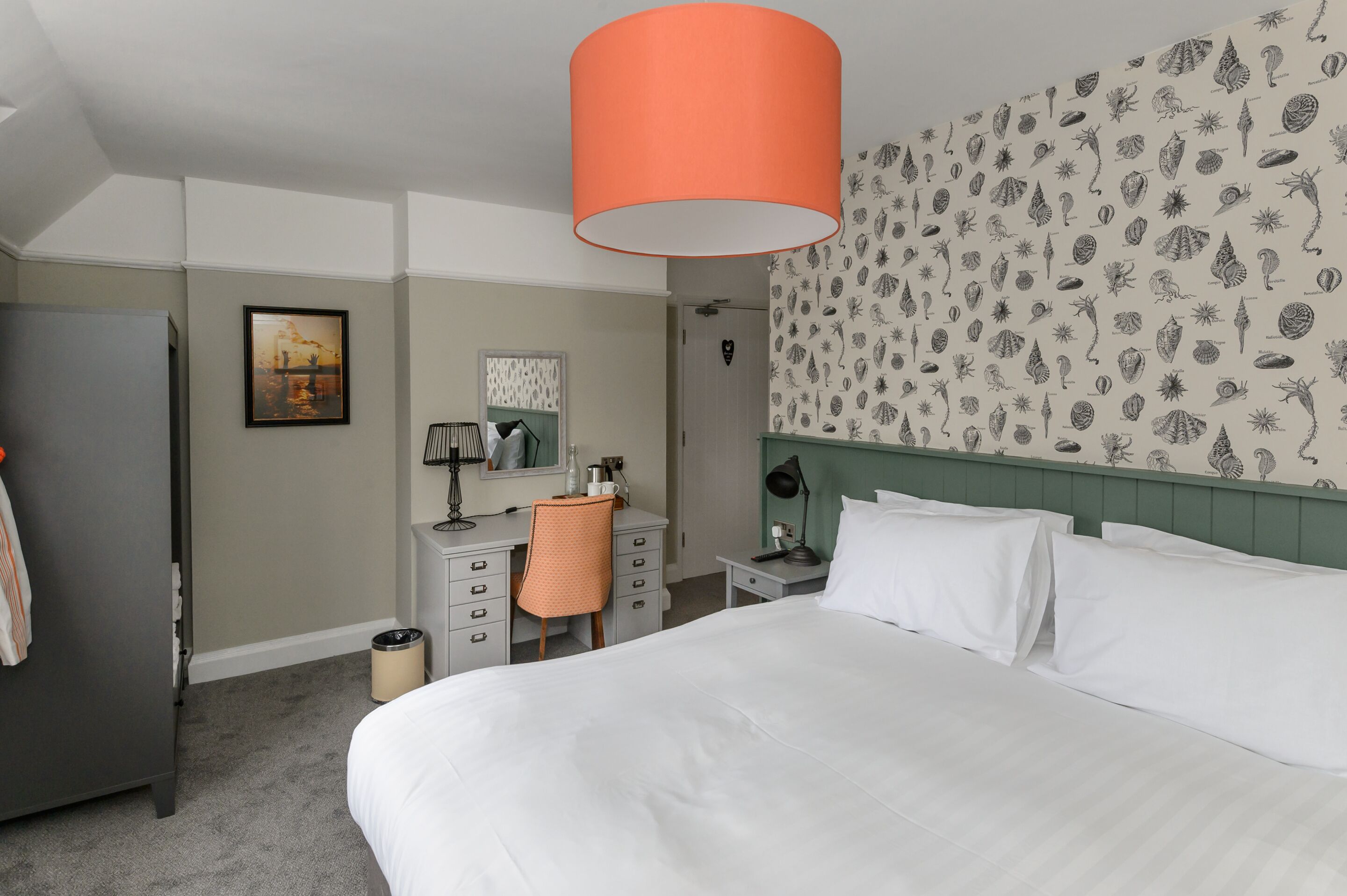 Habitación de hotel con una cama grande, un escritorio con silla, una mesita de noche, una lámpara y un panel decorado con un motivo de conchas.