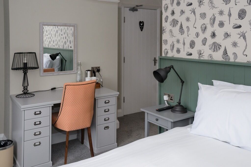 A bedroom with a bed, a desk with a chair, a nightstand, a lamp, and a wall with a marine life pattern.