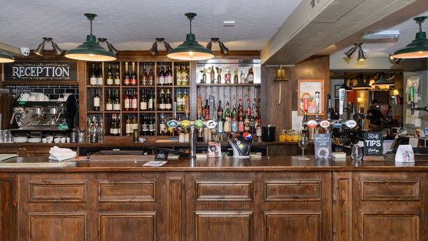 A bar area with wooden cabinets, a reception desk, and a variety of liquor bottles. - Balfour Arms (Sidmouth)
