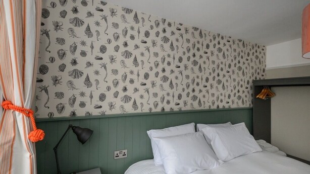 A hotel room with a bed, a nightstand with a lamp, and a wall with a leaf pattern. - Balfour Arms (Sidmouth)