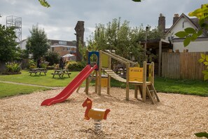 Un parque infantil con un tobogán y estructuras para escalar, rodeado por una valla de madera y una mesa de picnic.