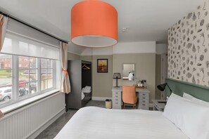 A hotel room with a bed, a desk, a chair, a lamp, a radiator, a window with curtains, and a wall with a leaf pattern. - Balfour Arms (Sidmouth)