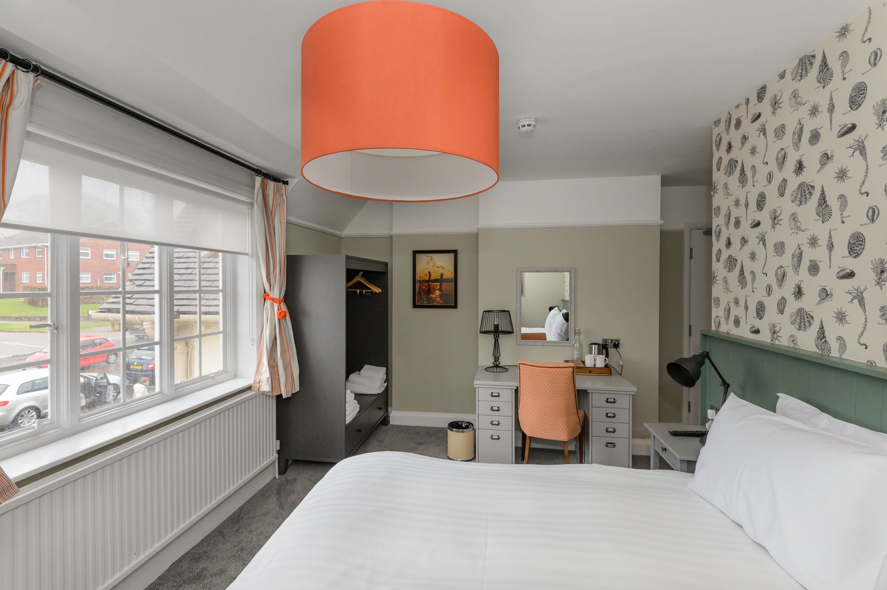 A hotel room with a bed, a desk, a chair, a radiator, a window with curtains, and a wall with a leaf pattern.
