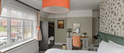 A hotel room with a bed, a desk, a chair, a radiator, a window with curtains, and a wall with a leaf pattern.