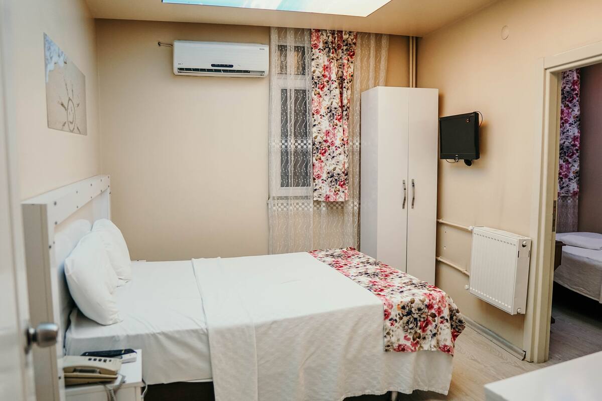 Ein Hotelzimmer mit zwei Betten, einem Kleiderschrank, einer Klimaanlage, einem Fernseher und einem Fenster mit blumigen Vorhängen.