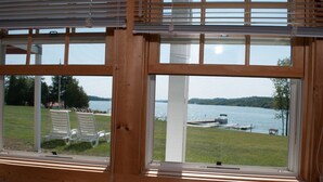 Une vue à travers une fenêtre à cadre en bois, donnant sur une zone herbeuse avec des chaises et un quai au bord d’un lac.