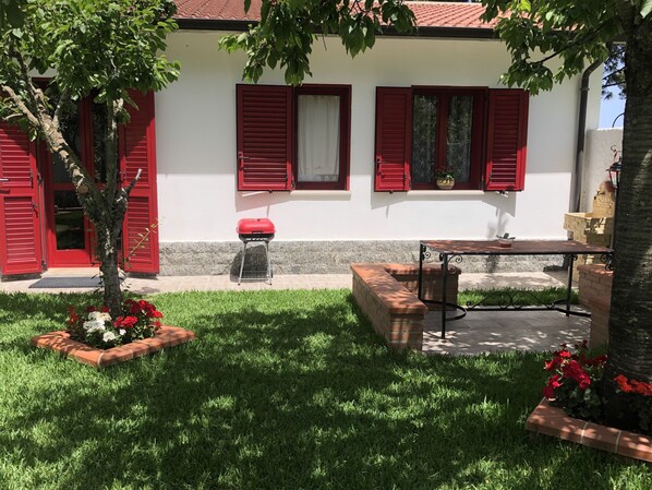 A white building with red shutters, a small garden with red flowers, and a table with a chair.