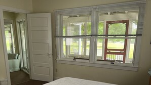 A bedroom with a large bed, a window with blinds, a door, and a view of a lawn and chairs outside. - Fountain Point - Gebhardt Cottage (Lake Leelanau)