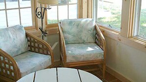 Une pièce avec deux fauteuils en osier et une petite table, de grandes fenêtres équipées de stores et une lampe sur une table d’appoint.