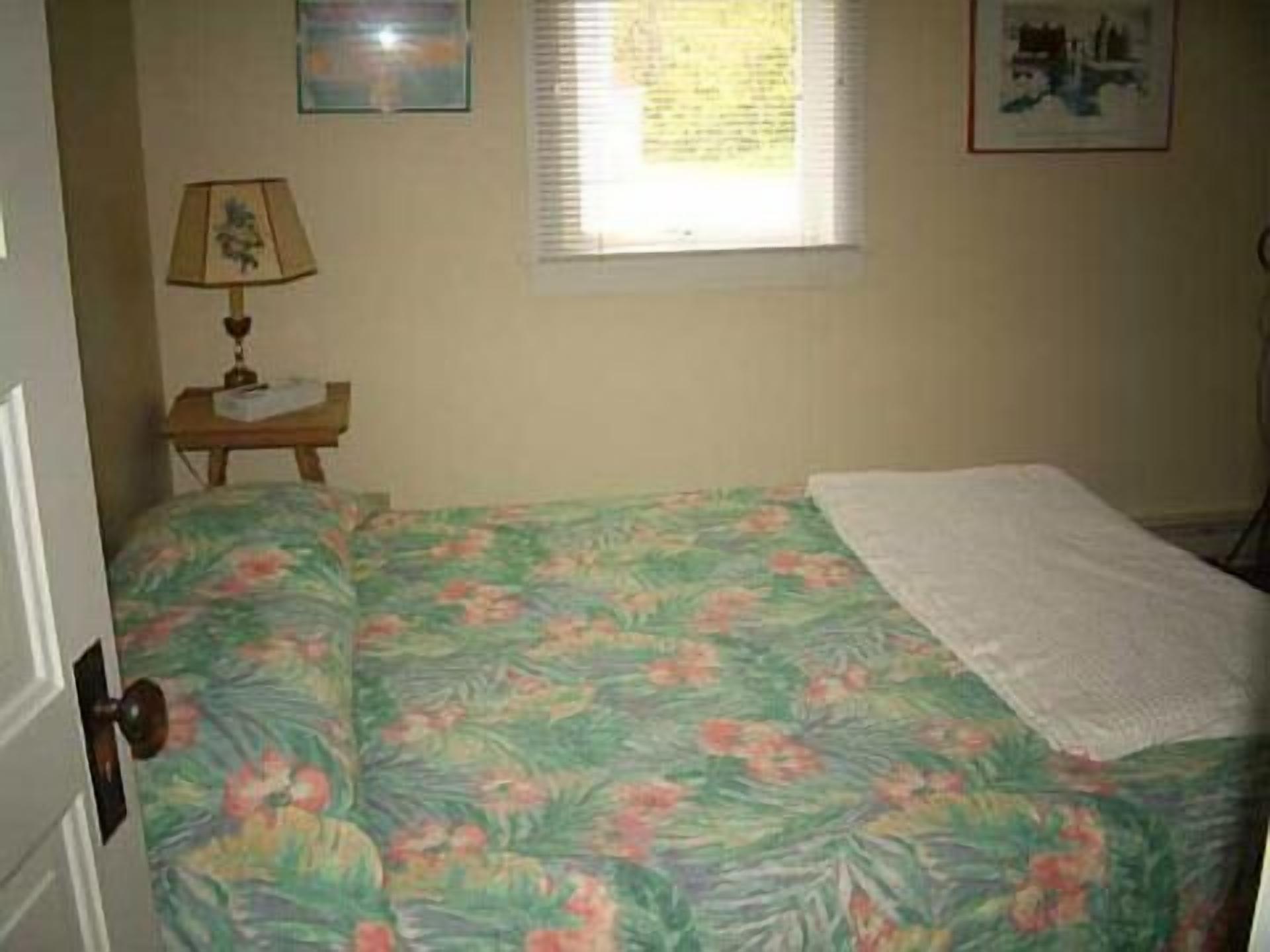 Un dormitorio con colcha estampada, una mesita de noche con lámpara y una ventana con persianas.