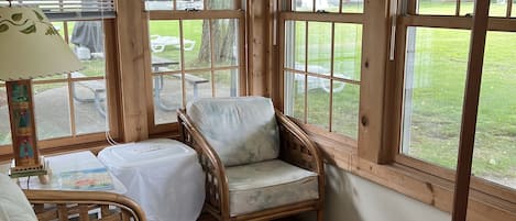Ein Wintergarten mit Holzrahmenfenstern, einem Korbsessel, einem kleinen Tisch und einer Sitzbank mit Auflage.