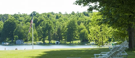 A row of white lounge chairs on a grassy area near a body of water, with trees and a flag in the background.