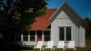 A white wooden building with a red roof, a porch with white chairs, and a large window. - Fountain Point - Shepherd Cottage (Lake Leelanau)
