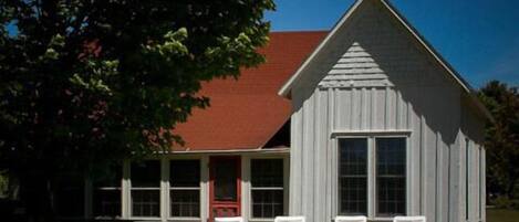 A white wooden building with a red roof, a porch with white chairs, and a large window.