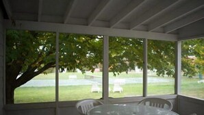 A covered patio with a round table and four chairs, large windows, and a tree outside. - Fountain Point - Cottage 17 (Lake Leelanau)