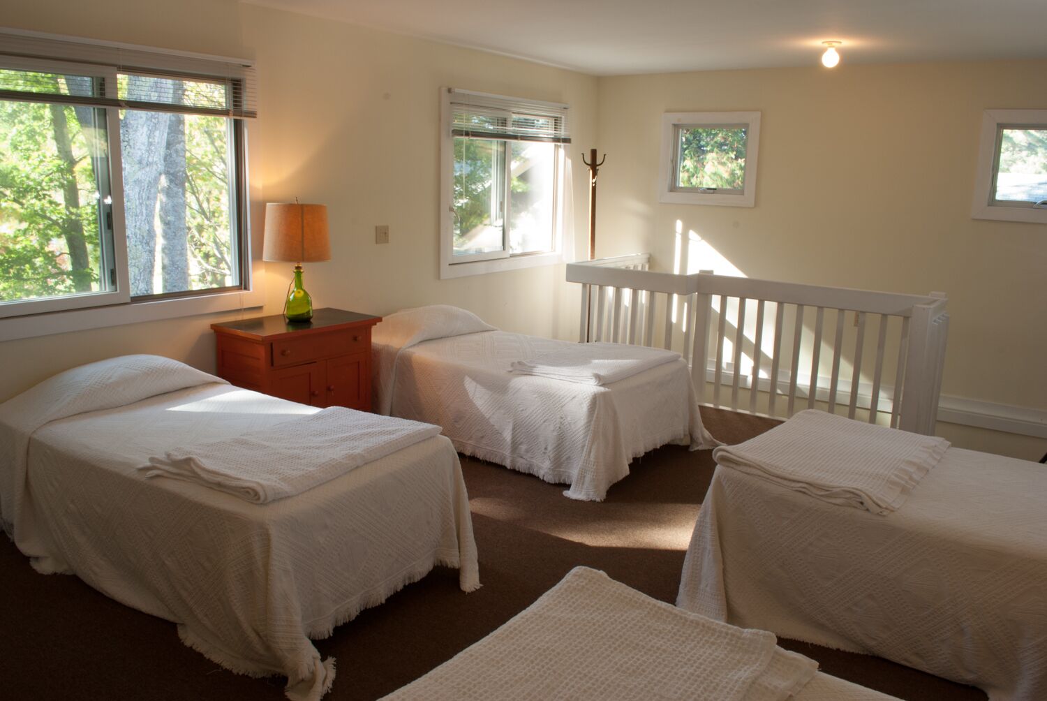 Un dormitorio con tres camas individuales, una mesita de noche, una lámpara y vistas a árboles por las ventanas.