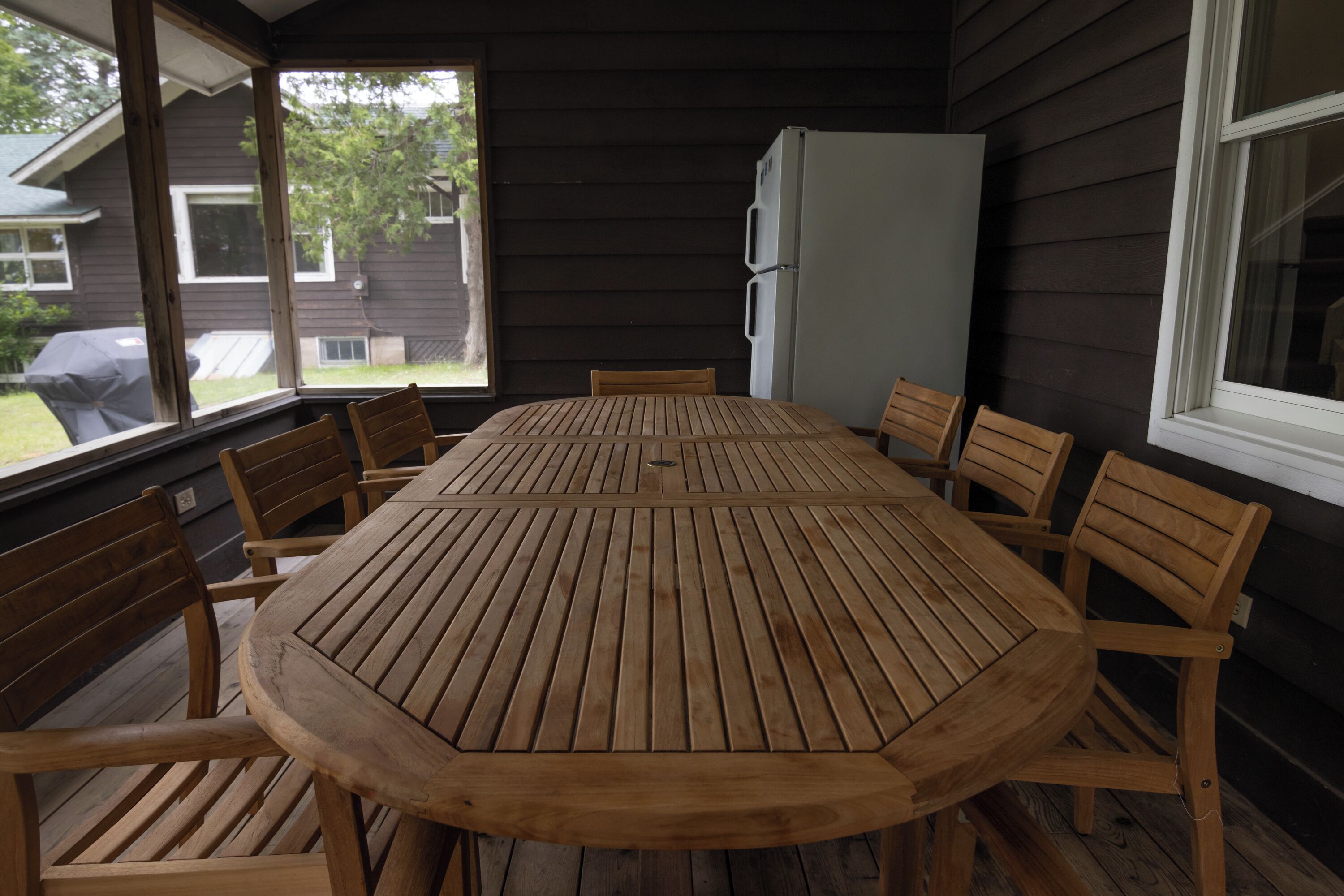A wooden table and chairs in a covered outdoor area with a view of a house and a refrigerator.