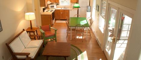 A compact kitchen with wooden cabinets, a microwave, and a sink. There is a dining area with a green table and chairs, and a sofa.
