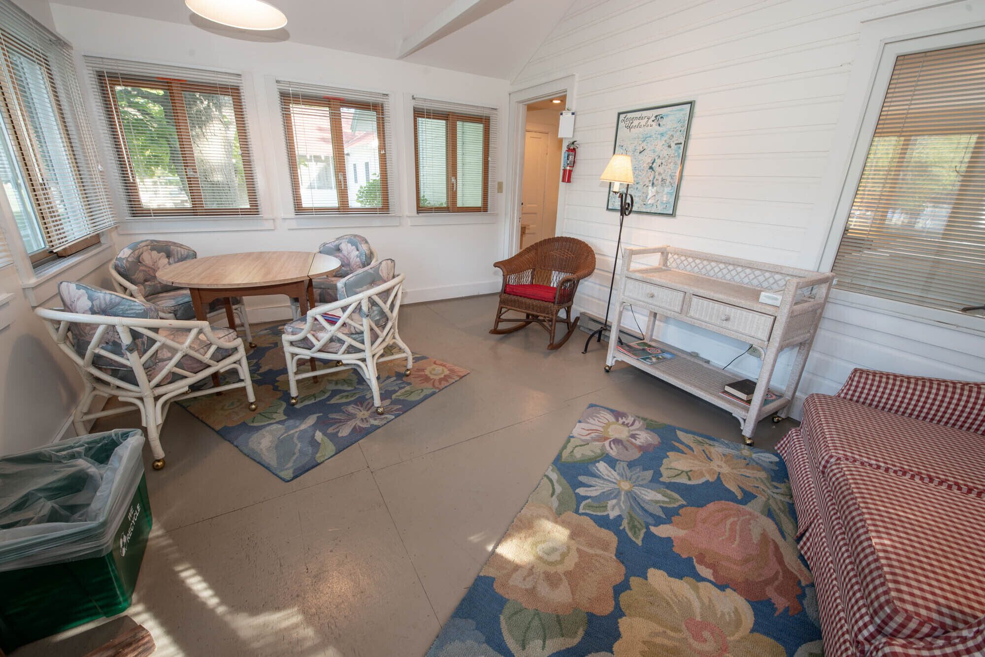 A room with a round table, wicker chairs, a rocking chair, a small white dresser, and a checkered sofa.
