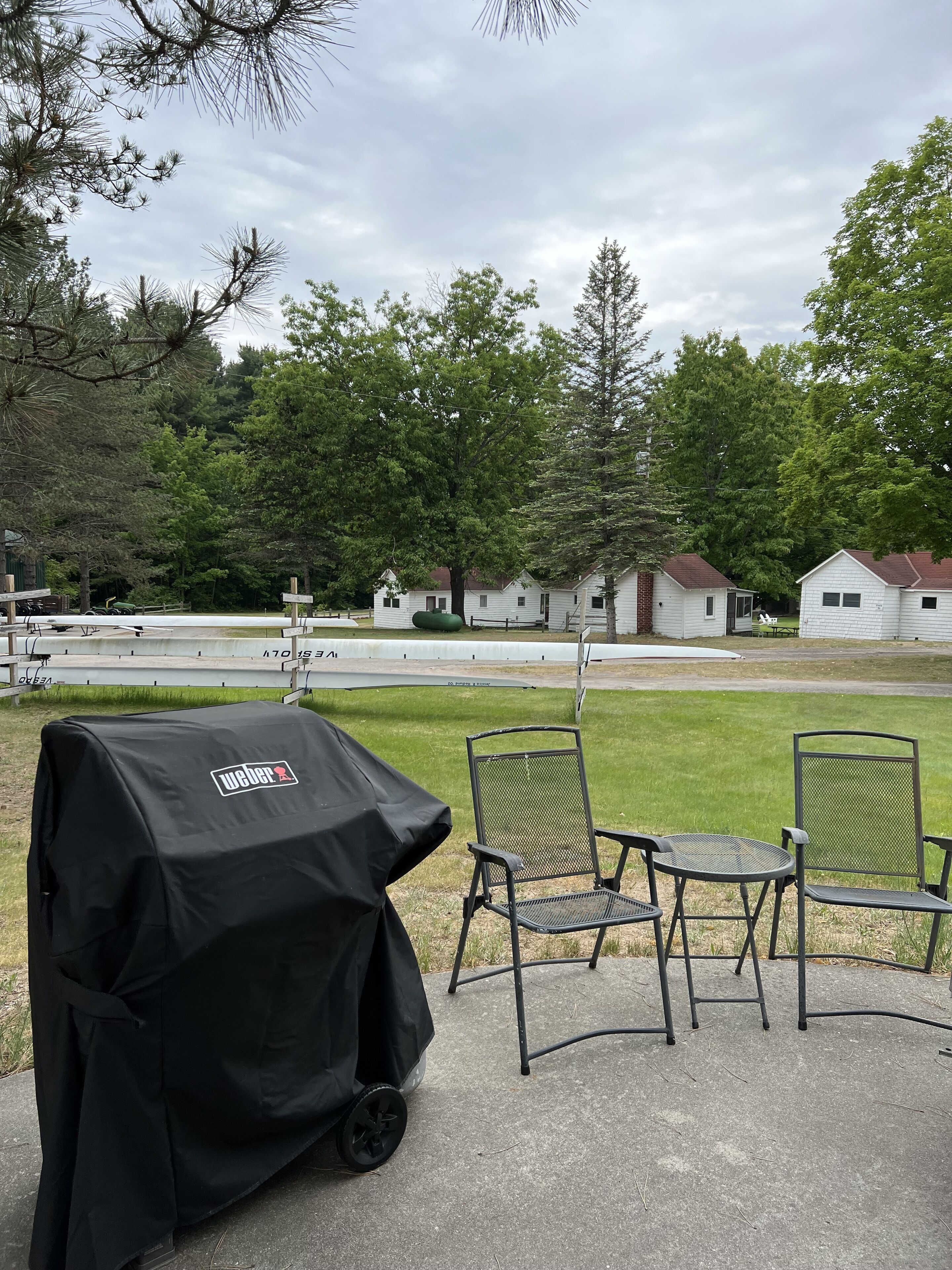 Een overdekte grill, twee metalen stoelen en een gazon met huizen op de achtergrond.