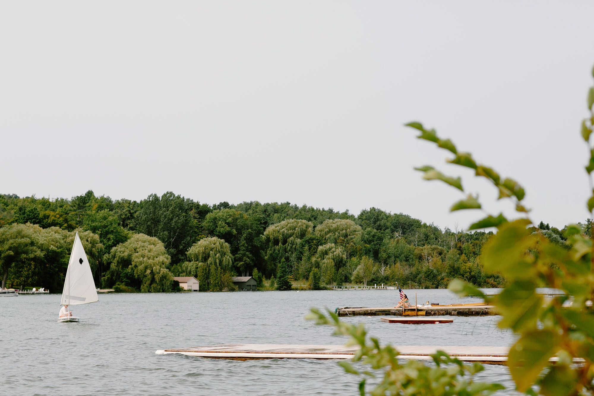 Un lac où l’on aperçoit un voilier et des bateaux amarrés, entouré d’une forêt dense et, au loin, de quelques bâtiments.