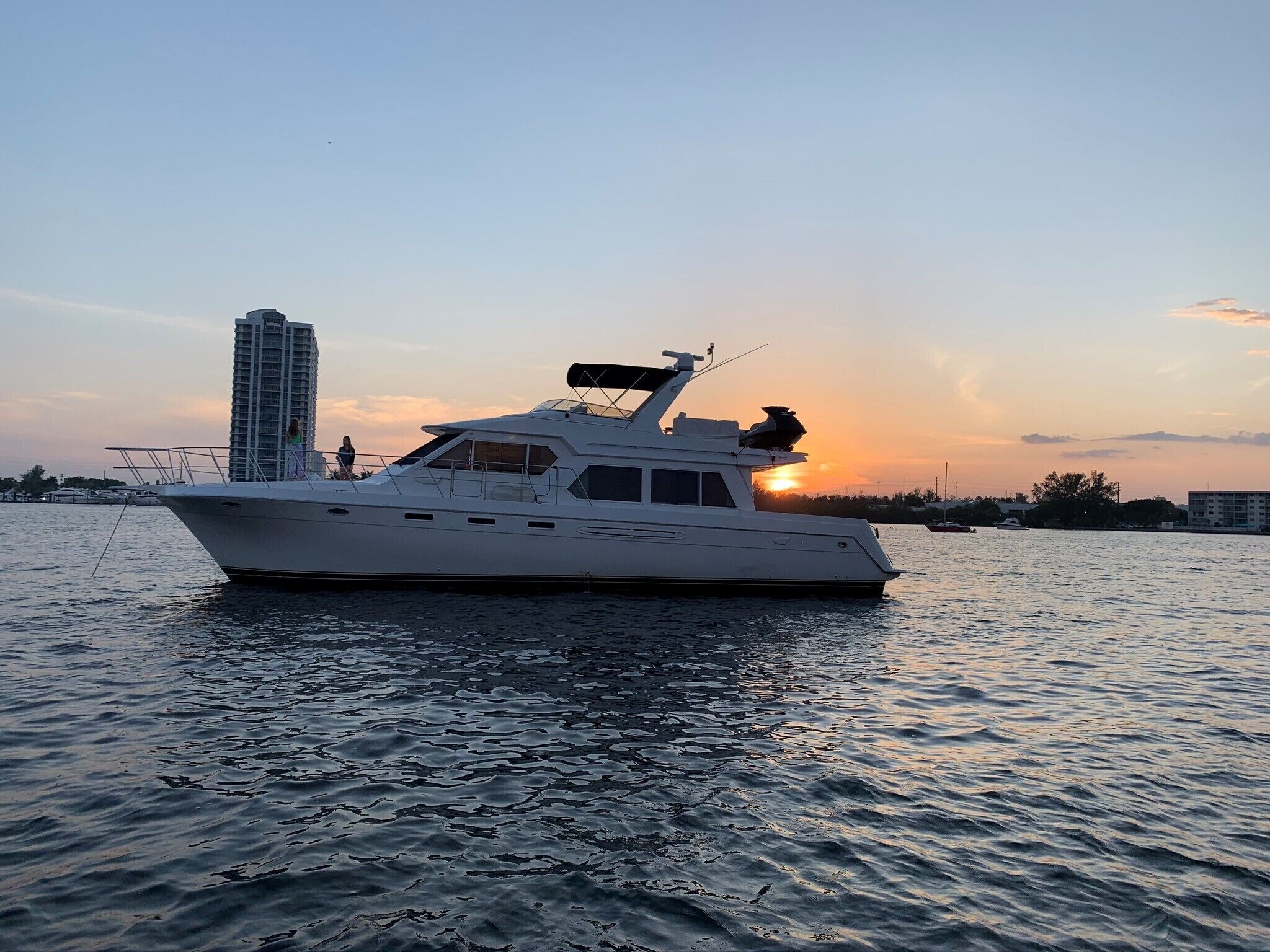 Un yate blanco con cabina y helipuerto en la cubierta, amarrado cerca de un edificio alto al atardecer.