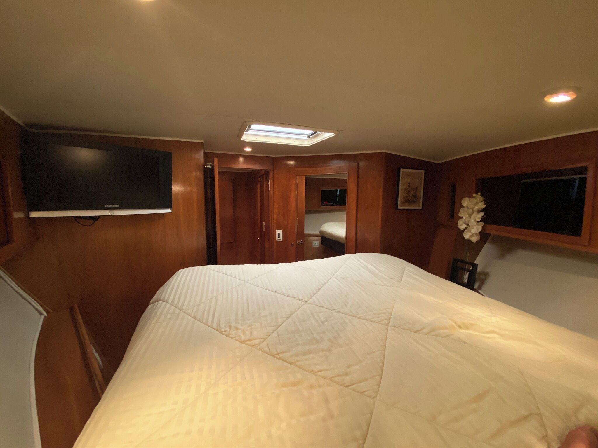 A bedroom with a bed, a television mounted on the wall, a ceiling light, and a door leading to another room.