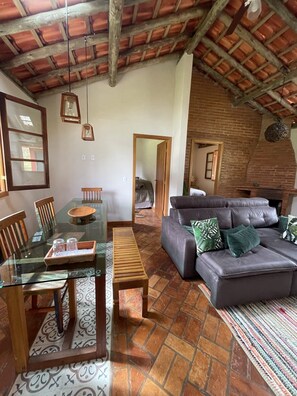 Uma sala de estar com uma mesa de jantar de vidro, cadeiras de madeira, um sofá cinza com almofadas verdes, um tapete estampado e uma lareira de tijolos.