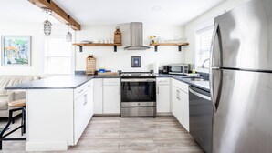 A modern kitchen with white cabinets, a stainless steel refrigerator, a built-in oven, and a microwave. There are wooden shelves above the counter and a hanging light fixture. - Contemporary  Elegance Meets Coastal decor (Kennebunk)