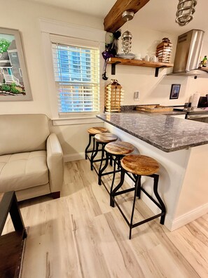 A kitchen with a marble countertop, wooden stools, a white sofa, and a window with blinds. - Contemporary  Elegance Meets Coastal decor (Kennebunk)