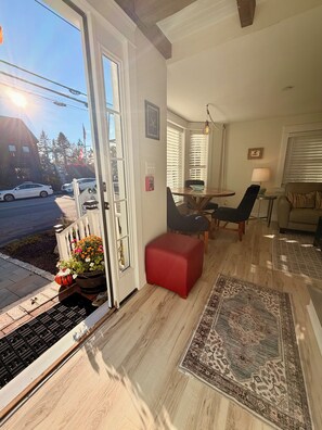 A modern living room with a wooden floor, a red ottoman, a dining table with chairs, a sofa, and a view of the street through a glass door. - Contemporary  Elegance Meets Coastal decor (Kennebunk)