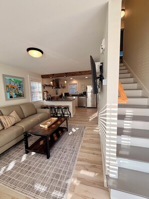 A modern living room with a sofa, coffee table, and a dining area with stools. A kitchen with a stove, refrigerator, and island is visible in the background. - Contemporary  Elegance Meets Coastal decor (Kennebunk)