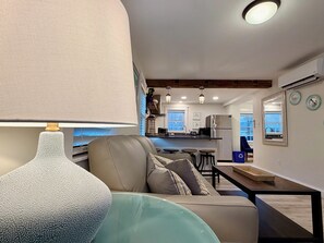 A modern living area with a sofa, a table, and a lamp. There is a kitchen in the background with a refrigerator and a sink. - Contemporary  Elegance Meets Coastal decor (Kennebunk)